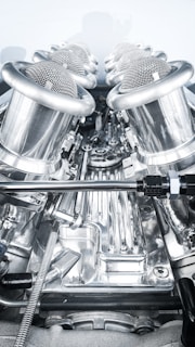A close-up view of a complex, polished metallic engine component, featuring a series of cylindrical, mesh-covered air intake manifolds. The metal surfaces are reflective and intricately designed, indicating high precision engineering.