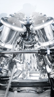 A close-up view of a complex, polished metallic engine component, featuring a series of cylindrical, mesh-covered air intake manifolds. The metal surfaces are reflective and intricately designed, indicating high precision engineering.