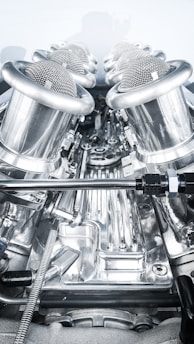 A close-up view of a complex, polished metallic engine component, featuring a series of cylindrical, mesh-covered air intake manifolds. The metal surfaces are reflective and intricately designed, indicating high precision engineering.