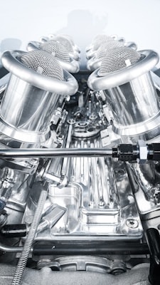 A close-up view of a complex, polished metallic engine component, featuring a series of cylindrical, mesh-covered air intake manifolds. The metal surfaces are reflective and intricately designed, indicating high precision engineering.