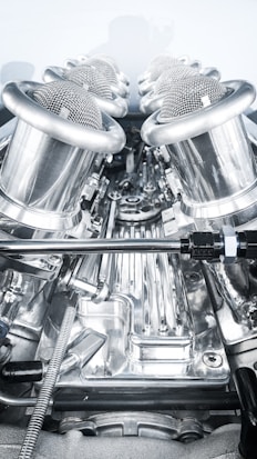 A close-up view of a complex, polished metallic engine component, featuring a series of cylindrical, mesh-covered air intake manifolds. The metal surfaces are reflective and intricately designed, indicating high precision engineering.