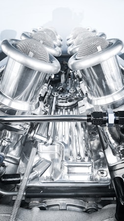A close-up view of a complex, polished metallic engine component, featuring a series of cylindrical, mesh-covered air intake manifolds. The metal surfaces are reflective and intricately designed, indicating high precision engineering.