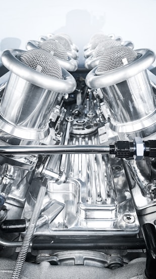 A close-up view of a complex, polished metallic engine component, featuring a series of cylindrical, mesh-covered air intake manifolds. The metal surfaces are reflective and intricately designed, indicating high precision engineering.