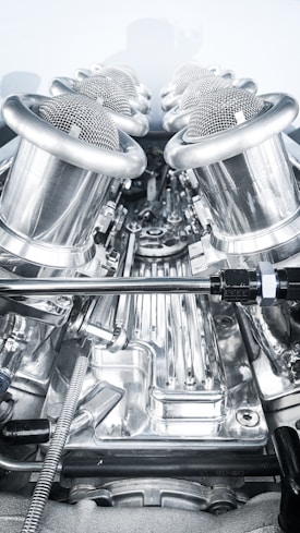 A close-up view of a complex, polished metallic engine component, featuring a series of cylindrical, mesh-covered air intake manifolds. The metal surfaces are reflective and intricately designed, indicating high precision engineering.