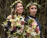 Two women are visible, both wearing flower crowns. They are each holding a large bouquet of flowers with various colors and textures, prominently featuring roses, berries, and different types of greenery. The bouquets are in focus, while the background is blurred, emphasizing the flowers.