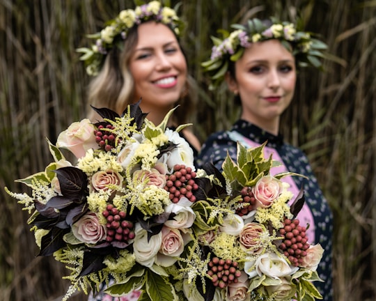Two women are visible, both wearing flower crowns. They are each holding a large bouquet of flowers with various colors and textures, prominently featuring roses, berries, and different types of greenery. The bouquets are in focus, while the background is blurred, emphasizing the flowers.