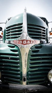 A close-up of the front grille of a vintage green Dodge truck, showcasing its distinctive chrome details and emblem. The polished metal reflects light, creating a contrast against the deep green paint.