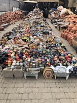A vibrant display of handmade pottery is arranged on the ground. The collection includes a wide variety of colorful ceramic dishes, bowls, and traditional tagine pots. Shelves and stacked arrays of earthenware line the sides, offering a rich assortment of designs and patterns. The setting appears to be an outdoor market with a mix of small and large clay products neatly organized for sale.