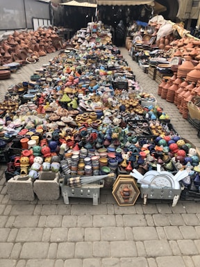 A vibrant display of handmade pottery is arranged on the ground. The collection includes a wide variety of colorful ceramic dishes, bowls, and traditional tagine pots. Shelves and stacked arrays of earthenware line the sides, offering a rich assortment of designs and patterns. The setting appears to be an outdoor market with a mix of small and large clay products neatly organized for sale.