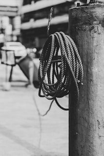 A coiled rope hangs on a metal post, with a blurred urban background featuring a parked car and buildings.