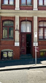 A brick building facade featuring a tall doorway with a dark brown door. Two symmetrical windows with red frames are positioned on either side of the door. Above the door, there is an arched window. A signpost in front indicates a no stopping zone. The building exhibits slight graffiti and weathering.