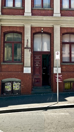 A brick building facade featuring a tall doorway with a dark brown door. Two symmetrical windows with red frames are positioned on either side of the door. Above the door, there is an arched window. A signpost in front indicates a no stopping zone. The building exhibits slight graffiti and weathering.