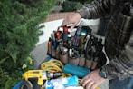 A person wearing a plaid shirt stands next to an organized tool bag filled with various tools such as screwdrivers, pliers, and a flashlight. A power drill, a can of cleaning spray, and coiled yellow cable are also visible. Lush greenery is present in the background.