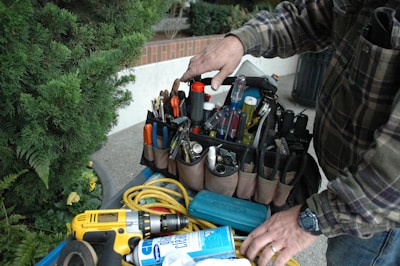 A person wearing a plaid shirt stands next to an organized tool bag filled with various tools such as screwdrivers, pliers, and a flashlight. A power drill, a can of cleaning spray, and coiled yellow cable are also visible. Lush greenery is present in the background.