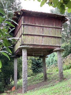 A traditional wooden granary elevated on stone pillars situated in a lush garden setting. The granary has a weathered appearance, composed of vertical wooden slats with a red tint on the aged wood. Surrounding the structure are green trees and foliage, indicating a rural or countryside location.
