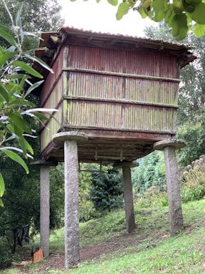 A traditional wooden granary elevated on stone pillars situated in a lush garden setting. The granary has a weathered appearance, composed of vertical wooden slats with a red tint on the aged wood. Surrounding the structure are green trees and foliage, indicating a rural or countryside location.