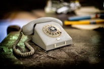 A vintage rotary dial telephone is placed on a worn table. The phone is beige, with a coiled cord on the left side. In the background, there are blurred objects including pens and papers, suggesting an office or study setting. The image has a nostalgic feel, with soft lighting highlighting the texture of both the phone and the table.