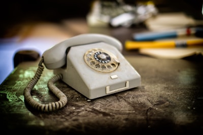 A vintage rotary dial telephone is placed on a worn table. The phone is beige, with a coiled cord on the left side. In the background, there are blurred objects including pens and papers, suggesting an office or study setting. The image has a nostalgic feel, with soft lighting highlighting the texture of both the phone and the table.