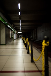 A dimly lit underground subway platform with tiled flooring and a long row of overhead lights stretching into the distance. Yellow chains are strung between black posts along the edge of the platform, with green and white signage visible on the left side.