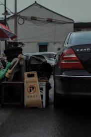 A black car is parked next to a pile of miscellaneous items including stacked tires, a green and white tent fabric, and a black plastic bag. A 'No Parking' sign with text in both English and Chinese is propped against a wooden stool. The background features a white building with a tiled roof and visible wires overhead.