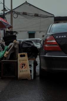 A black car is parked next to a pile of miscellaneous items including stacked tires, a green and white tent fabric, and a black plastic bag. A 'No Parking' sign with text in both English and Chinese is propped against a wooden stool. The background features a white building with a tiled roof and visible wires overhead.