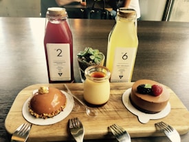 A wooden serving board on a table holds various desserts and beverages. There are two bottles of juice, one red and one yellow, with labels indicating health benefits. A small potted succulent plant is placed between them. In front, there is a caramel-glazed pastry with a nut crust, a jar of pudding with caramel topping, and a chocolate mousse cake garnished with fresh berries. Three forks are positioned nearby.