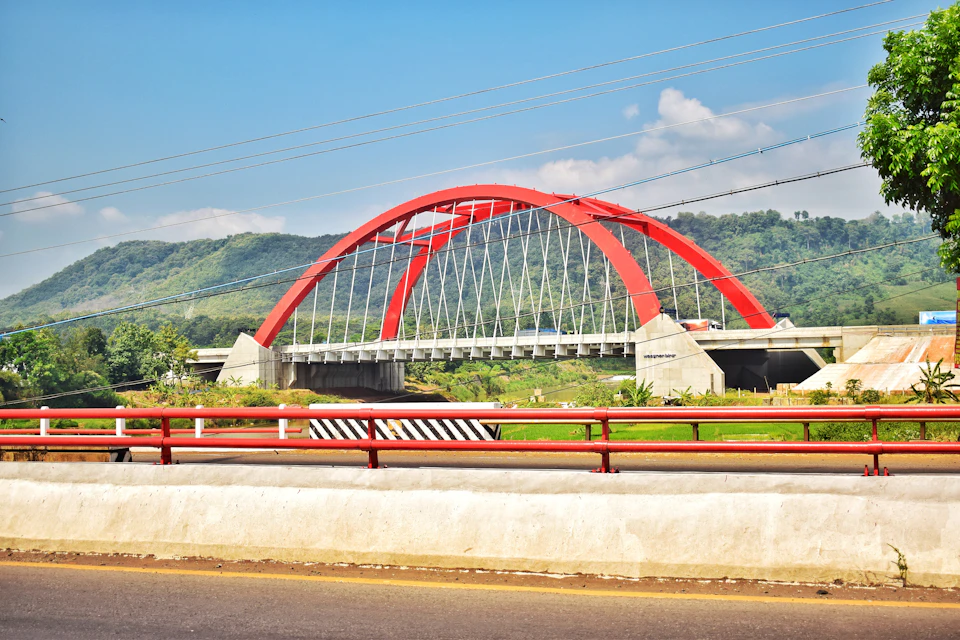 The image depicts a striking red arch bridge spanning across a river, with its curved structure and vibrant color contrasting against the serene backdrop of lush green trees and rolling hills. The bridge is situated on an elevated road, providing a clear view of the surrounding landscape. Power lines can be seen in the distance, indicating that this picturesque scene is likely located near a populated area or town.

The perspective of the image is from the opposite side of the river, looking towards the bridge. This viewpoint offers a unique vantage point to appreciate both the natural beauty and human ingenuity behind the construction of such an impressive structure.