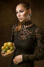 A woman wearing a black lace dress holds a bowl filled with lemons and limes. Her hair is tied back, and she wears ornate jewelry, including earrings and a necklace. The background is dark, emphasizing her contemplative expression and poised posture.