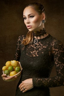 A woman wearing a black lace dress holds a bowl filled with lemons and limes. Her hair is tied back, and she wears ornate jewelry, including earrings and a necklace. The background is dark, emphasizing her contemplative expression and poised posture.