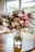 A bouquet of pink and white flowers arranged in a glass jar, tagged with a label that reads 'Bridesmaid'. The bouquet is composed of roses, greenery, and other floral elements, sitting on a wooden table with a blurred background indicating an indoor setting.