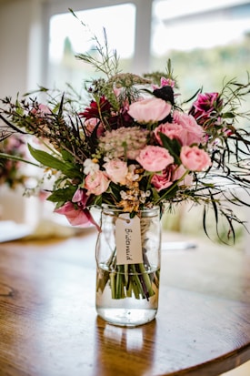 A bouquet of pink and white flowers arranged in a glass jar, tagged with a label that reads 'Bridesmaid'. The bouquet is composed of roses, greenery, and other floral elements, sitting on a wooden table with a blurred background indicating an indoor setting.