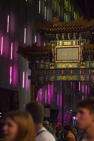 A decorative archway with intricate patterns and Chinese characters is prominently featured, surrounded by a night scene with people walking by. The background is illuminated with vibrant neon lights in shades of pink and green, creating a lively urban atmosphere.