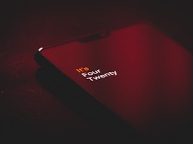 A smartphone lays on a dark surface, emitting a faint red glow. The screen displays the text 'It's Four Twenty' in white and orange letters, creating a contrast against the dark surroundings.