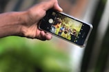 A person is holding a smartphone horizontally, capturing a photo of a group of people outdoors. The phone screen displays the group of friends standing together in a grassy area, suggesting they are socializing and having fun.
