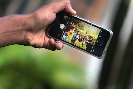 A person is holding a smartphone horizontally, capturing a photo of a group of people outdoors. The phone screen displays the group of friends standing together in a grassy area, suggesting they are socializing and having fun.