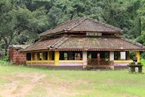 A small, rustic building with a traditional tiled roof and yellow walls is situated in a lush, green forest setting. The building appears to be a temple with a sign in a local script above its entrance. The surrounding area is grassy and slightly overgrown, enhancing the natural, tranquil ambiance.