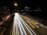 Long exposure photograph of a busy urban street at night, showcasing bright, colorful light trails from passing traffic. The scene is illuminated by street lights, with buildings and trees visible in the background.