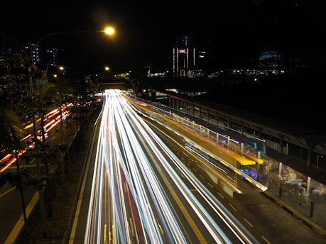 Long exposure photograph of a busy urban street at night, showcasing bright, colorful light trails from passing traffic. The scene is illuminated by street lights, with buildings and trees visible in the background.