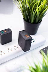 A neatly arranged workspace featuring a black charging block plugged into a white power strip. Surrounding the power strip are black and white electronic accessories, including another charging block and a set of earbuds. A black potted plant with vibrant green leaves adds a touch of nature to the modern setting, while a smartphone lies nearby on the white surface.