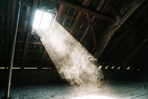 Sunlight streams through a small window in the roof of an attic, casting a soft, illuminating beam that highlights the dust particles floating in the air. The wooden beams of the ceiling are visible, and the space has an aged, rustic feel.