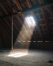 A dusty attic with wooden beams and a small window in the slanted roof. Sunlight streams through the window, highlighting particles floating in the air. The floor is old and weathered, creating a rustic atmosphere.