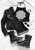 A stylish flat lay of fashion items including a black leather jacket, a black halter top with a white intricate mandala design, black denim shorts, lace-up black combat boots with studs, a pair of black sunglasses, and black bangles. The items are arranged on a white cloth background.