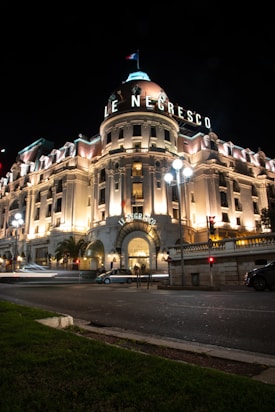 An elegant historical hotel building with illuminated arched entrance, classical architecture, and a rooftop dome under a starless night sky. The facade is adorned with intricate details and a sign displaying 'Le Negresco'. Palm trees and streetlights cast a warm glow across the pavement, while cars create light trails on the road in front of the building.