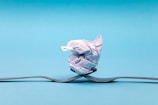 A crumpled piece of paper balanced on two intersecting forks against a solid light blue background. The forks are positioned in a way that they appear to be supporting the paper ball, creating a sense of balance and symmetry.