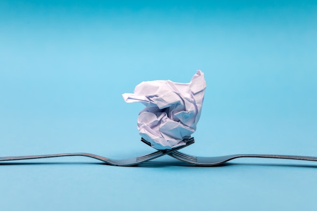 A crumpled piece of paper balanced on two intersecting forks against a solid light blue background. The forks are positioned in a way that they appear to be supporting the paper ball, creating a sense of balance and symmetry.