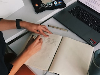 A person happily writing song lyrics on a notebook with a laptop open showing a lyrics generator interface.
