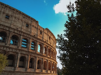 An ancient stone amphitheater with multiple arches is prominently featured, partially obscured by lush green trees on the right side. The building's facade showcases several tiers of arched openings, with some areas under restoration, indicated by scaffolding inside the arches. The sky is predominantly blue with a few scattered clouds, creating a contrast with the aged texture of the stone structure.