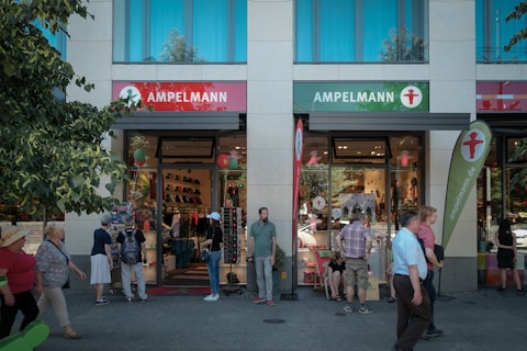 A bustling street scene with people walking near a shop named Ampelmann. The storefront has large glass windows displaying merchandise, and several people are visible both inside and out. The building has tall windows and is flanked by trees on one side. Various flags and signs with the Ampelmann logo, a silhouette of a walking figure, are prominently displayed.