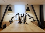 A modern radio studio setup with a long wooden table and multiple microphones mounted on adjustable arms. Two large pendant lights hang overhead, providing focused lighting. The walls are a neutral color, creating a clean and professional atmosphere. The letters 'ACI RADIO' are visible on the wall, indicating the studio's branding.