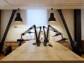A modern radio studio setup with a long wooden table and multiple microphones mounted on adjustable arms. Two large pendant lights hang overhead, providing focused lighting. The walls are a neutral color, creating a clean and professional atmosphere. The letters 'ACI RADIO' are visible on the wall, indicating the studio's branding.